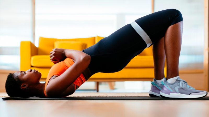 Fitness athlete performing banded glute bridge exercise with resistance loop band on yoga mat in sunlit apartment studio